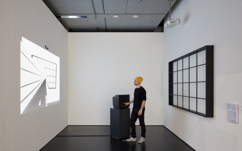 Das Foto zeigt eine Projektion zur linken und ein gerastertes Bild auf der rechten Wand. In der Mitte hält ein Mann mit Mütze seine Hand in einen Röhrenfernsehen.
