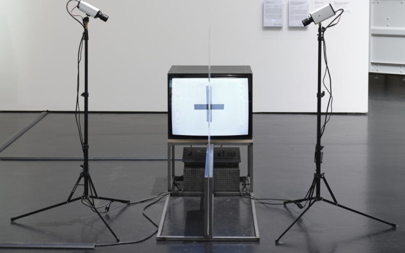 Das Foto zeigt einen Röhrenbildschirm der von zwei Kameras umrahmt wird. Die Kameras sind auf zwei Ständern montiert. Mittig zum Bildschirm ist auf einem Metallgestell eine Plexiglas Platte aufgestellt. Ein Kreuz leuchtet aus dem Rohrenbildschirm.  