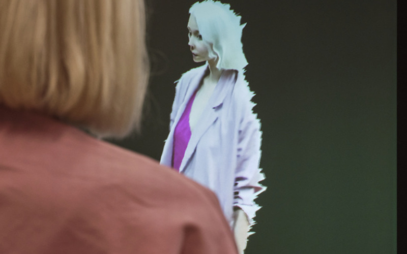 Im Vordergrund des Fotos sieht man eine hellblonde Frau mit kurzen Haaren und einer rosafarbenen Jacke. Vor ihr ist sie als Avatar auf einem Bildschirm reproduziert. Die Besucherin steht ihrem digitalen Code gegenüber.