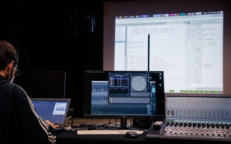 Live Coding Performance: Eine Person ist von hinten zu sehen, vor ihr ist ein Computerbildschirm und eine große Leinwand, auf der ein Programmiercode zu sehen ist.