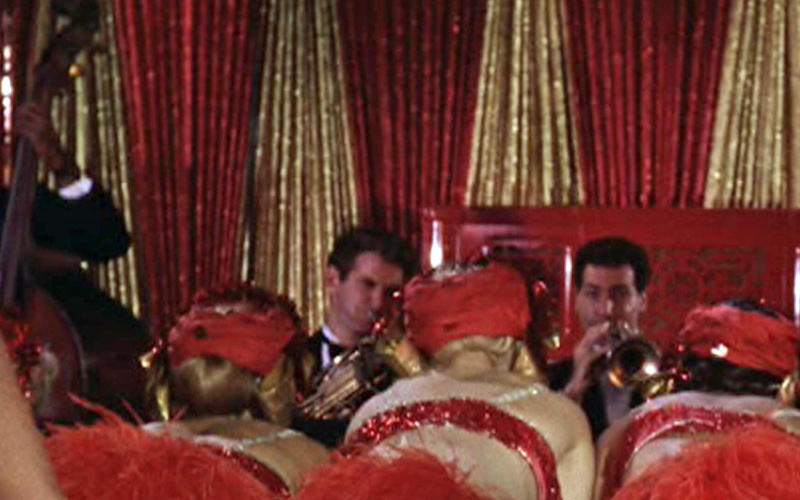 Two trumpet players are seen in front of a golden and red curtain. In the audience sit three women with red headdresses