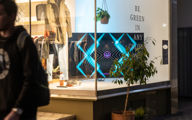 Die interaktive Lichtinstallation wird auf einem großen Bildschirm gezeigt, welcher sich im Schaufenster eines Bekleidungsgeschäftes befindet.
