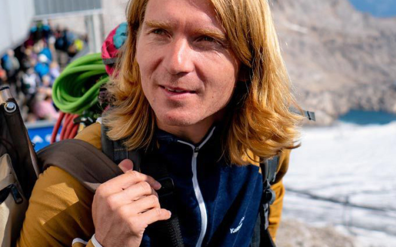 The picture shows the artist Markus jeschanuig. He is pictured outside and seems to be on a hike on a mountain top. He wears open shoulder-length blond hair and looks left out of the picture.