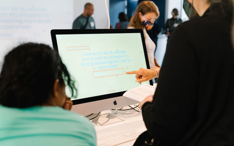 Auf dem Bild zu sehen ist ein Monitor auf dem zwei halb im Bild befindenden Menshen etwas gezeigt wird. Auf dem Monitor ist der Schriftzug "How might we're imagine our Future reationship to Berlin Brandenburg Water Ecosystem" zu lesen