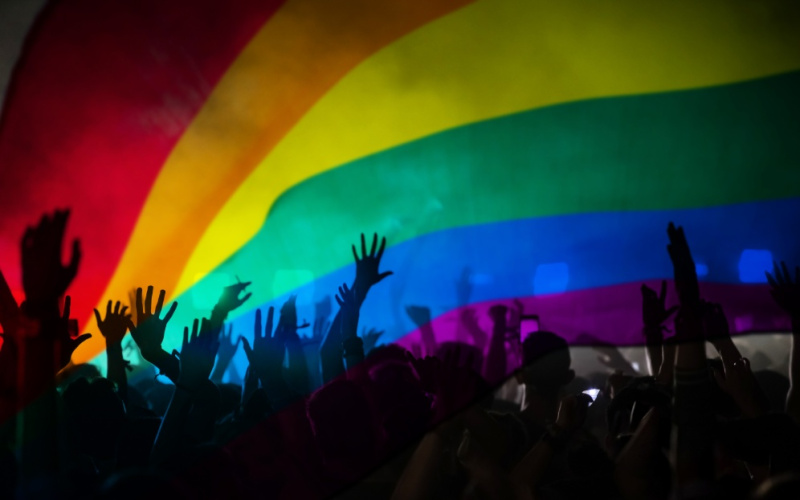 Zu sehen sind Hände vor einer Regenbogenflagge.