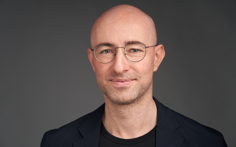 The image depicts the portrait of the media artist Alexander Peterhänsel. He is wearing glasses and is smiling into the camera.
