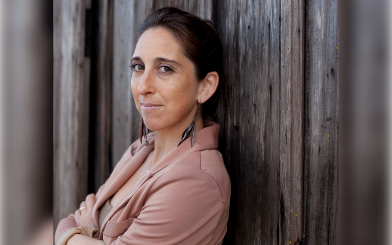 A woman with is leanig against a wooden background with her arms crossed. She is wearing a dusty pink jacket and earrings. Her hair and eyes are brown and she is smiling into the camera. 