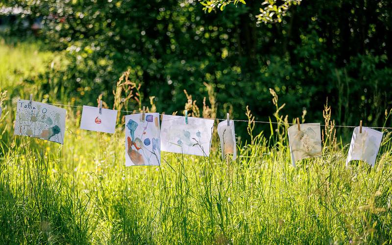 Zu sehen sind Bilder, die an einer Leine über hohem Gras auf einer Wiese hängen.