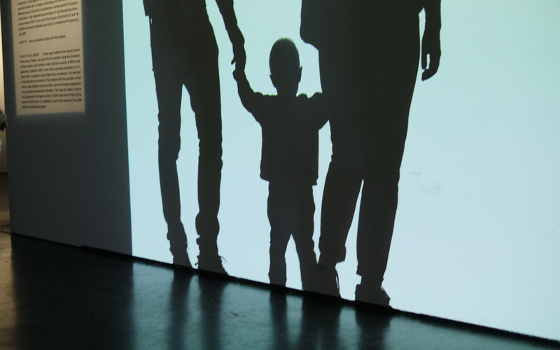 Schatten eines Kindes, das die Hände zweier Erwachsenen hält vor weißem Hintergrund.