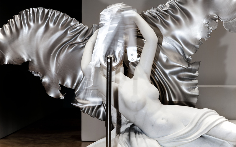 Vor dem Kopf einer klassischen Marmorstatue mit silbernen Flügeln bewegt sich unscharf eine silberne Masse.