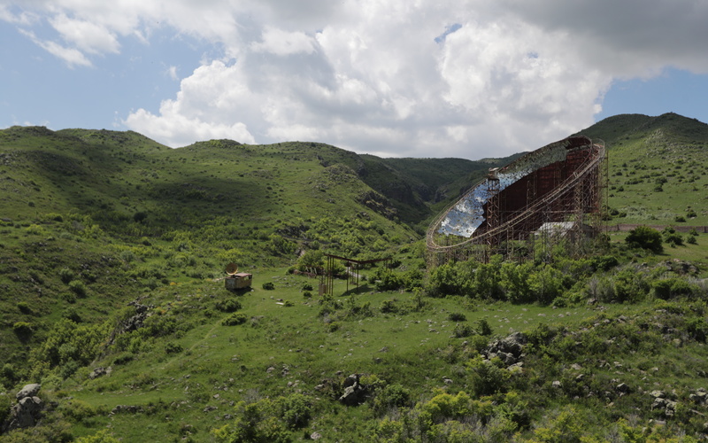 In a green, hilly landscape, a large curved metal structure with reflective surfaces stands beneath a cloudy sky.