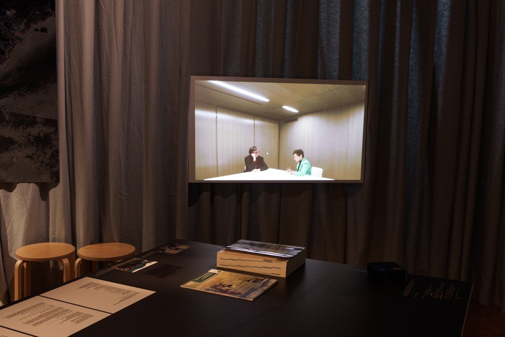 Tisch mit Exponaten und Hockern im Vordergrund, ein Bildschirm im Hintergrund.