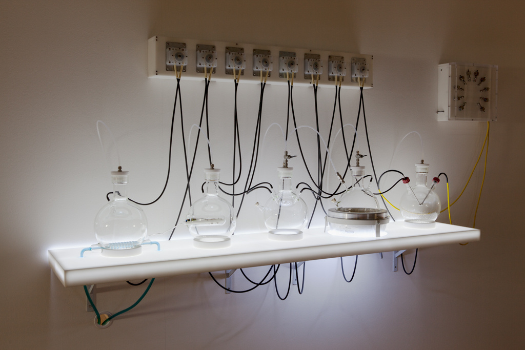 Experimenteller, laborartiger Versuchsaufbau auf einer leuchtenden Wandablage.