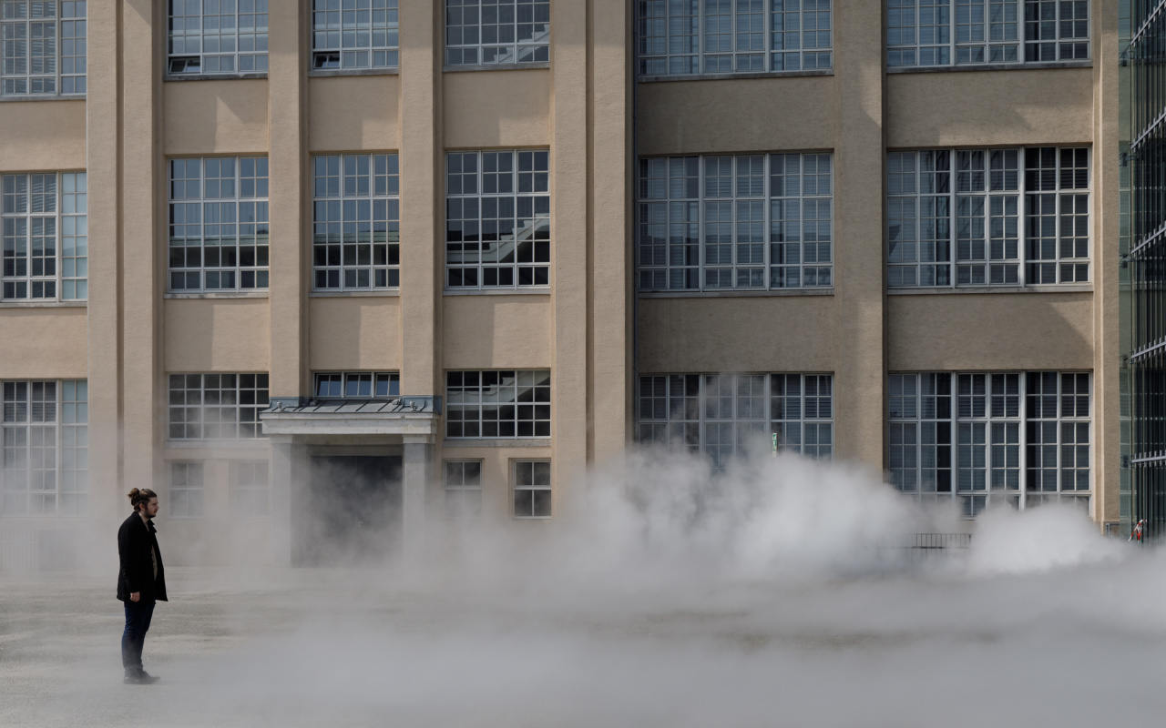 Das Foto zeigt einen Mann ummantelt von der Nebelskulptur der japanischen Künstlerin Fujiko Nakaya. Im Hintergrund ist die Fassade der alten Munitionsfabrik zu sehen.