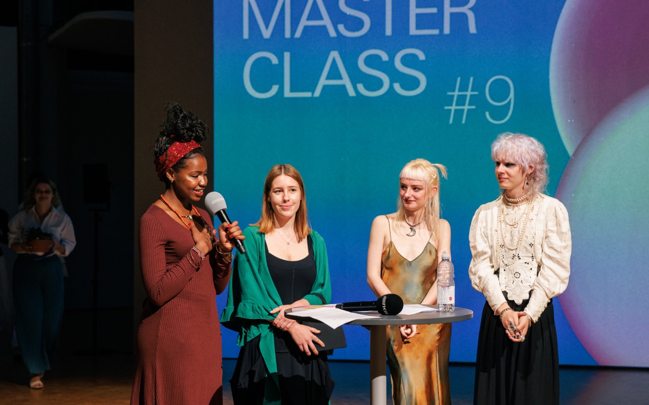 Vier Personen auf der Bühne der [MASTERCLASS] #9-Eröffnung. Die Person ganz links hält in der einen Hand ein Mikrofon und hat die andere Hand auf ihr Herz gelegt.