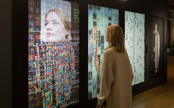A woman stands in front of a wall with several screens and digital representations