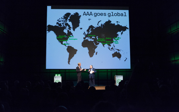 Zwei Männer auf der Bühne. Im Hintergrund eine Weltkarte mit »AAA goes global«