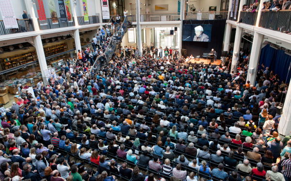 Vor einer Bühne, auf der ein Mann referiert, sitzen viele Menschen auf Stühlen