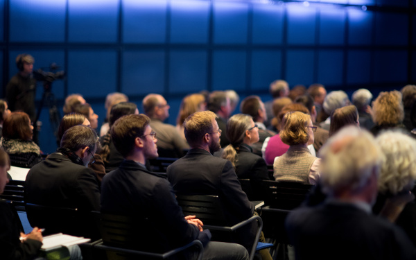Viele Menschen sitzen in einem Saal. Man sieht sie von hinten.
