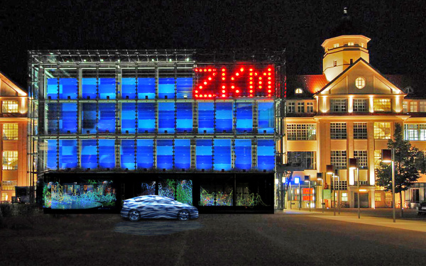 A car stands in front of the blue cube. Top right in big red glowing letters: ZKM