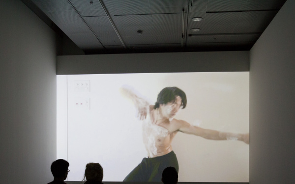 Three persons sit in front of a screen on which a man, wrapped in cling film, can be seen