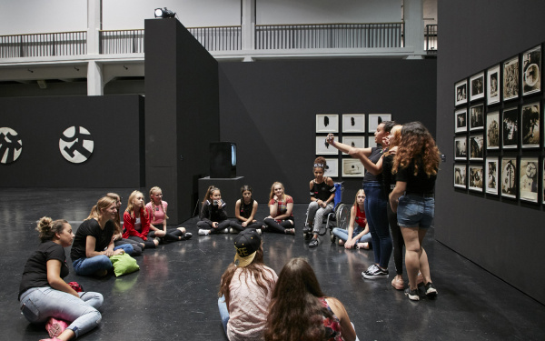 Eine Gruppe von Mädchen sitzt im Kreis in einer Ausstellung. Drei von ihnen machen ein Selfie vor einem Kunstwerk.