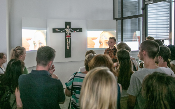 On the wall hangs a cross with a skeleton, as a triptych two videos are shown next to it. A crowd stands in front of the artwork.