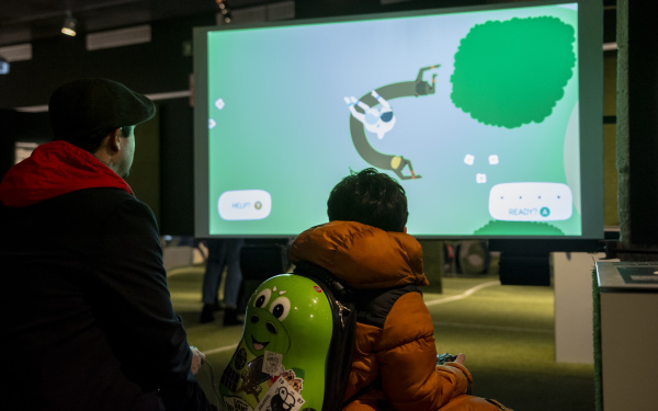 Ein Mann und ein kleiner Junge sind mit dem Rücken zum Betrachter gewandt. Der Junge hat einen birnenförmigen Rucksack an, der einen Computerspielcharakter verkörpert. Im hinteren Teil des Bildes ist ein Bildschirm mit Spiel zu sehen, was gespielt wird. 