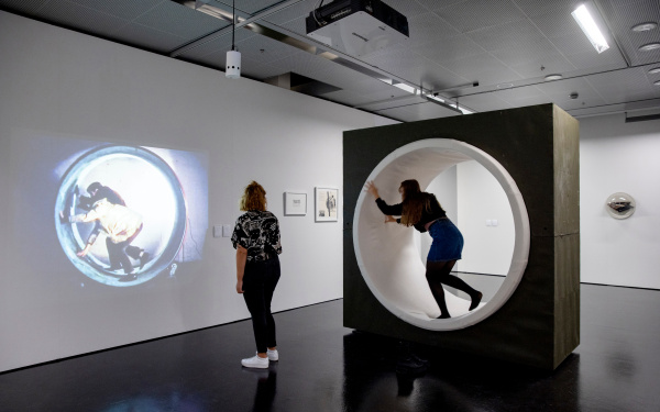 Zwei junge Frauen sind im Ausstellungsraum. Eine betrachtet eine Projektion eines künstlerischen Films an einer Wand und die andere befindet sich in einer Lauftolle.