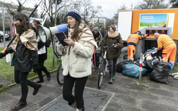 Eine Gruppe von Menschen gehen in einer Prozession mit Gartenwerkzeugen, Fahrrädern und Megafon. 