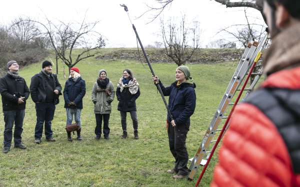 Eine Gruppe von Menschen stehen auf einer Wiese und in der Mitte hält eine Frau eine Baumsäge.