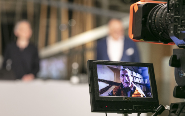 Im Vordergrund ist ein Video-Kamera-Display mit Bruno Latour drauf zu sehen. Hinter dem Display sind eine Frau und Peter Weibel verschwommen zu erkennen.