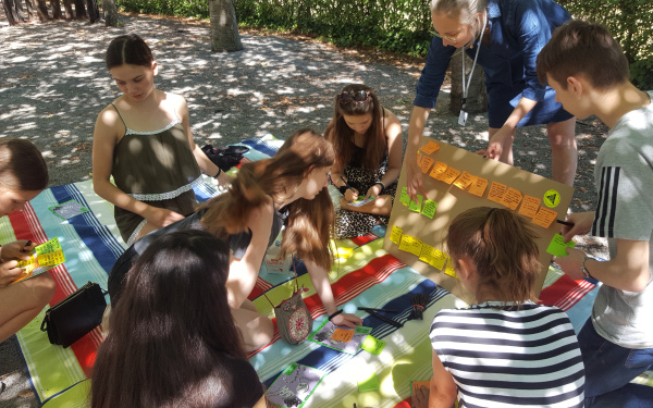 Eine Gruppe von Jugendlichen sitzt auf einer Picknick-Decke im Freien und hören einer Frau zu, die mit ihnen spricht.