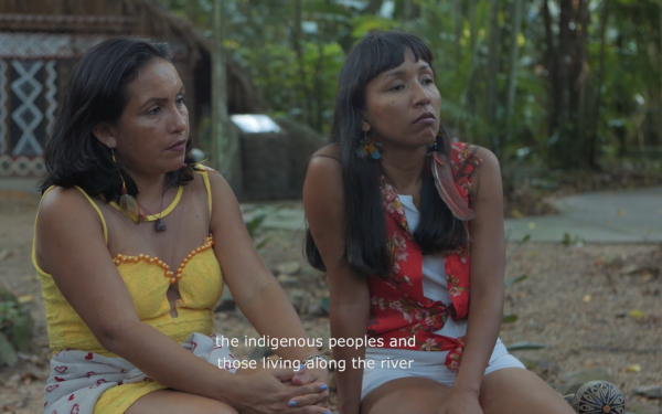 Two women can be seen, who are interested in hearing someone outside the picture. Around them it is green and rural.