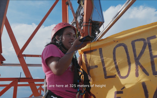 The picture shows a woman on a steel frame holding a large banner in one hand and a smartphone in the other, which she talks into. You can read: we are at 325 meters of height. 