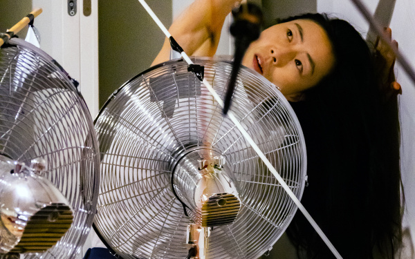 Das Foto zeigt eine koreanische Performerin die nach links gebeugt ihren Arm über ihren Kopf ausstreckt, während vor ihr ein großer silberfarbener Ventilator steht.