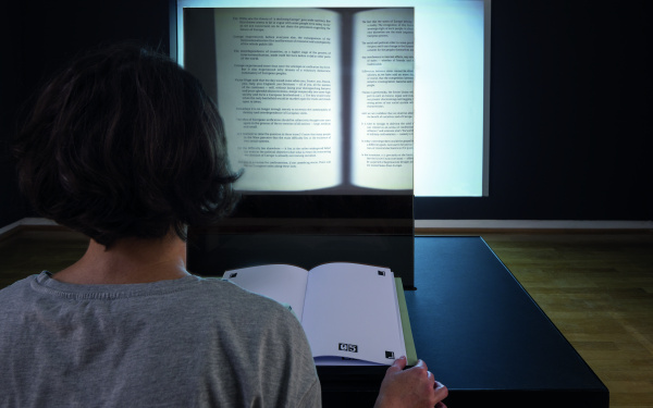 Zu sehen ist der Hinterkopf einer Person, vor ihr liegt ein aufgeschlagenes Buch. An der Wand gegenüber ist eine Projektion eines aufgeschlagenen Buches.