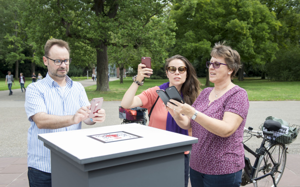 Leute mit Smartphone stehen vor einem Sockel im Freien.