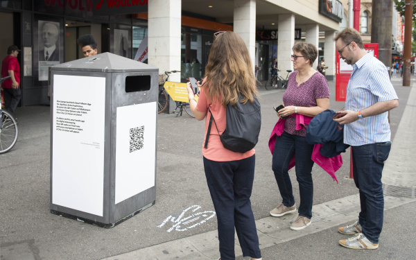 Leute mit Smartphone stehen vor einem Mülleimer mit QR-Code