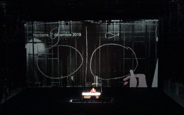 Bruno Latour is sitting on a stage behind a desk. Behind him is a large screen showing two large circles, with a long, vertical line between them.