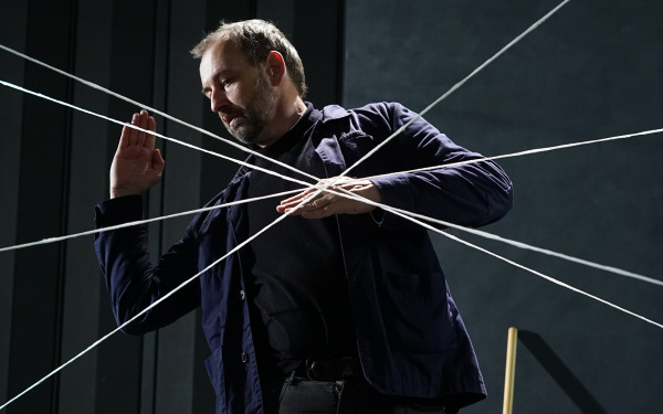 Mehrer weiße Fäden ziehen sich über die Fotografie wo ein Mann mit ernstem Gesicht eine strenge Pose mit angewinkelten Armen hält.