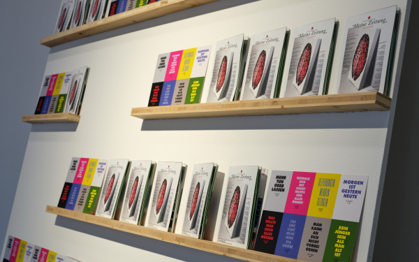 Zu sehen ist eine Wand, an welcher fünf Regale aus einzelnen Bretter befestigt wurden. Auf jedem der Regale stehen Flyer der Ausstellung. 