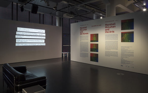 Im Vordergrund auf der linken Seite steht ein schwarzes Sofa. Hinten links steht eine Stellwand mit Text-Projektion. Hinten rechts eine mit Text und Bildern bedruckte.