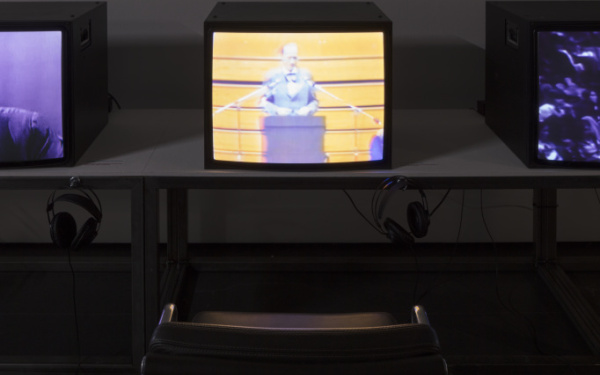 Nahaufnahme einer Videoinstallation. Fernseher auf Tisch mit Kopfhörern und davor ein schwarzes Stuhl.