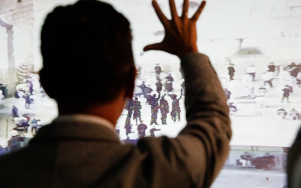 A picture can be seen on a wall on which shadowy figures raise their hands to salute. In front of it stands a man, his back to the observer and also raising his hand.