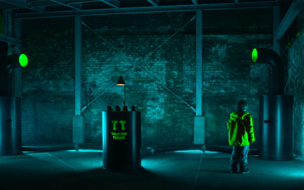 Ein blau erleuchteter Raum mit einem Kind, das die »Talking-Tubes«-Installation betrachtet: ein Steuerungspult zwischen zwei großen Röhren