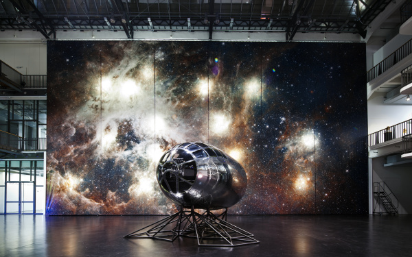 Zu sehen ist ein kleines, silbernes Gestellt vor einer großen Leinwand mit dem Motiv einer Galaxie, das an eine Raketenspitze erinnert. 