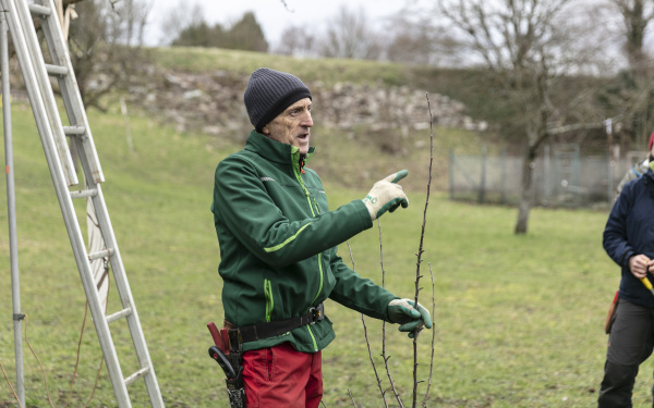 Ein Mann in Outdoor-Kleidung erklärt etwas und hält einen Ast in der Hand.
