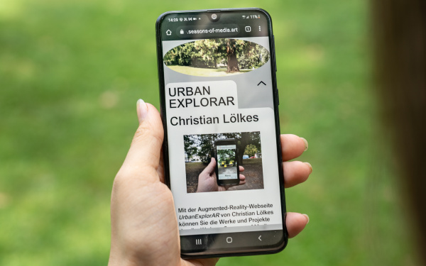 Auf dem Bild ist eine Hand zu sehen, die ein Smartphone hält, auf dem das Werk »UrbanExplorAR« zu sehen ist.