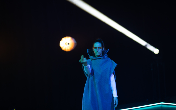 Zu sehen ist eine Person in blauer Kutte vor schwarzem Hintergrund. Ein weißer Lichtstrahl wieso ein runder Lichtkegel sind hinter ihr zu erkennen.